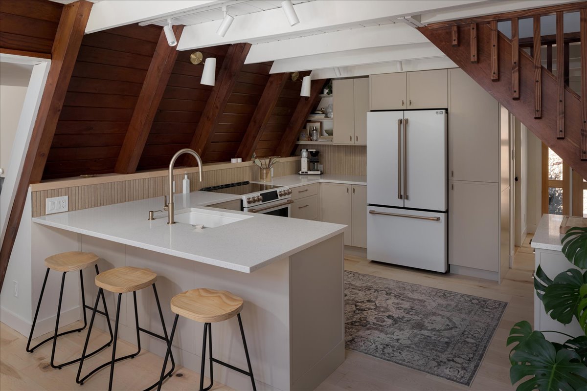 Opened A-frame kitchen with white island, stools, refrigerator, and stair railing